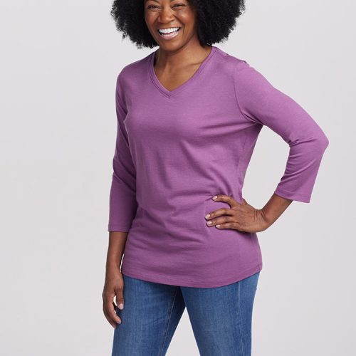 A woman with curly hair smiles while striking a modern chic pose. She is wearing the elegant Elena VNeck from Woolx in the color Dewberry, crafted from luxurious merino wool, paired with blue jeans. Her left hand rests on her hip as she stands against a plain white backdrop.