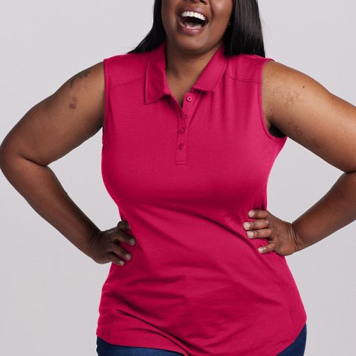 A woman wearing a Rosie Sleeveless Polo from Woolx in Viva Magenta and blue jeans stands with her hands on her hips, smiling broadly against a plain, light gray background.