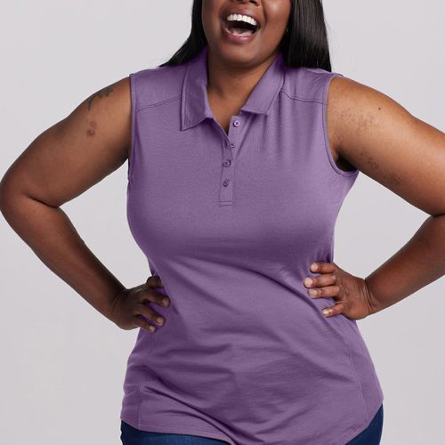 A woman in a Montana Grape Rosie Sleeveless Polo by Woolx and blue jeans is laughing with her hands on her hips. She stands against a plain gray background, showcasing effortless style and joy.