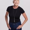 An older woman with short white hair smiles confidently in a black Gracie Bodysuit OLD by Woolx and dark jeans, making it ideal for a chic summer wardrobe. A long necklace adds flair as she stands with her hands in her pockets against a plain white background.