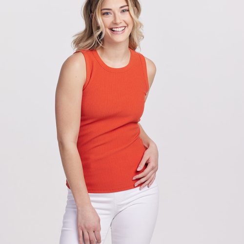 A person with long, wavy hair smiles at the camera, wearing a Raegan Ribbed Tank Top in Summer Fig and white pants. The sleeveless top, from Woolx, enhances their style against the plain white background.