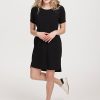 A person with curly hair is smiling while standing in the Lexie Dress & Cover-up OLD by Woolx, a black knee-length dress, paired with white sneakers against a plain background.