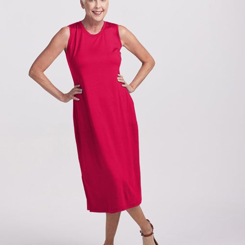 An older woman with short white hair poses confidently in the Cassie Dress by Woolx, a sleeveless Viva Magenta midi dress featuring side pockets, paired with brown sandals. She stands against a plain white background, her hands on her hips.