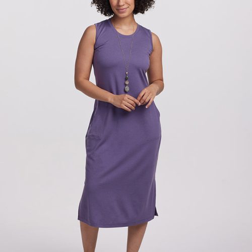 A person with curly hair wears the sleeveless Cassie Dress in Montana Grape from Woolx, paired with sandals. They pose against a plain white background, smiling gently while holding a long necklace with a pendant.