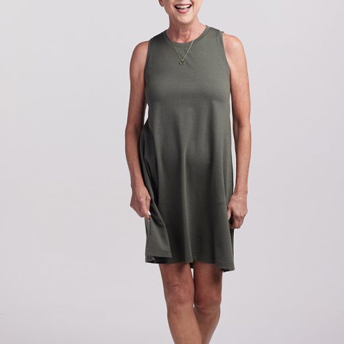 An older adult with short white hair smiles while standing against a plain background. They are wearing a sleeveless, knee-length Clara dress in Deep Fern from Woolx and brown sandals. They hold the stylish dress at the sides, showcasing its simple elegance.