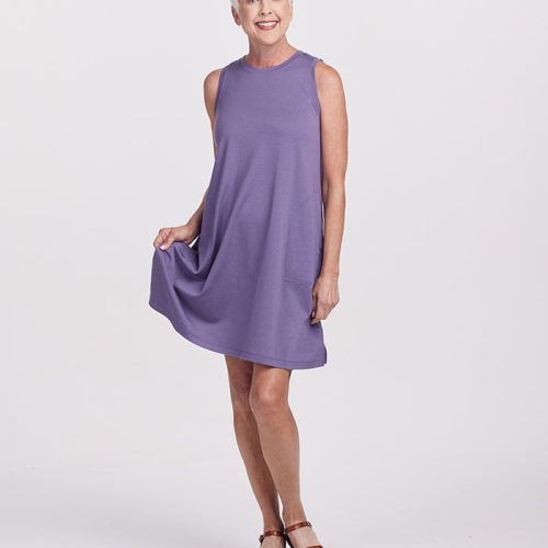 A person with short white hair smiles while wearing the sleeveless stylish Clara Dress in Montana Grape from Woolx and brown sandals. They are standing on one foot, holding the dress with one hand. The background is a plain light gray.