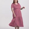 A person wearing a Mesa Rose, short-sleeve Lucia Dress from Woolx poses against a plain white background. With brown hair and a smile, they gently lift part of the tiered dress to showcase its elegance. They complete the look with brown sandals.