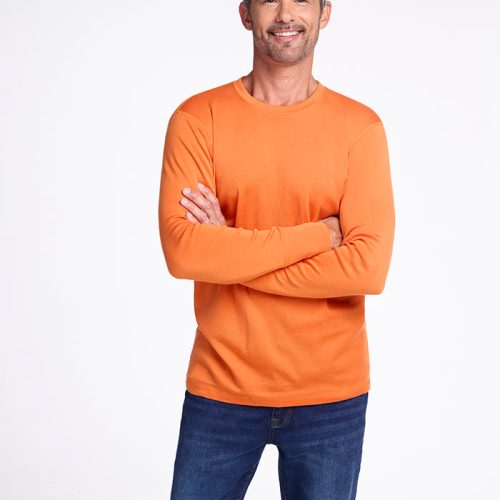 A man with short gray hair and a beard is standing with his arms crossed. He is wearing the Explorer Base Layer - Harvest Pumpkin from Woolx, paired with blue jeans. He is smiling against a plain white background. | Matthew is 6', wearing a size L