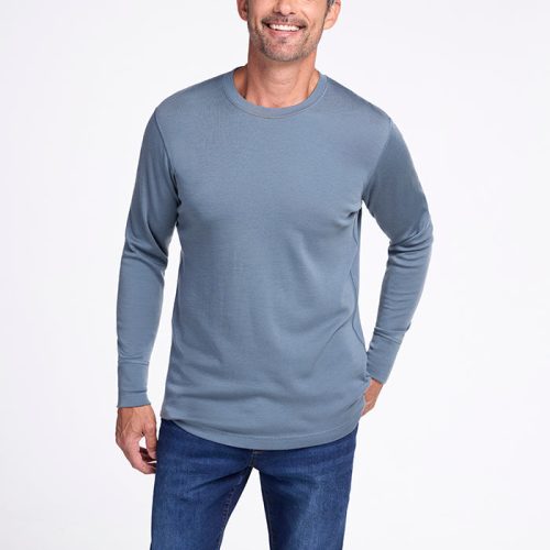 A man with short, gray hair and a beard is smiling and posing against a plain light background. He is wearing the Woolx Explorer Base Layer - Stormy Weather, a long-sleeved, midweight performance shirt in light blue, along with dark blue jeans. One hand is in his pocket while the other rests by his side, ready for his next outdoor adventure. | Matthew is 6', wearing a size L