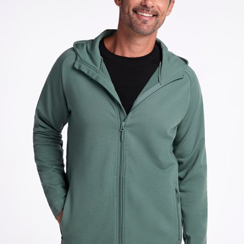 A smiling man with gray hair is wearing a Duck Green, breathable Merino wool Boulder Hoodie from Woolx over a black t-shirt. He has his left hand in his hoodie pocket. The background is plain and white.