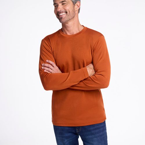 A smiling man with short gray hair stands confidently, arms crossed, in a cinnamon stick-colored Easton Crew Neck Shirt by Woolx, layered over a Merino base layer and paired with blue jeans. The background is plain white.