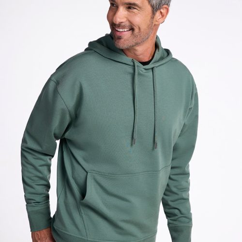 A man with short, gray hair is smiling while wearing the Chase Hoodie - Duck Green from Woolx and jeans. The adventure sweatshirt features drawstrings and a front pocket. He is standing against a plain, white background and looking slightly to his right. | Matthew is 6', wearing a size L