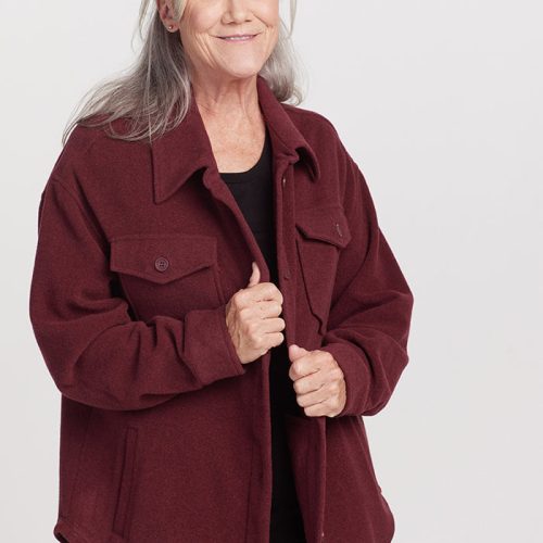 An older woman with long gray hair smiles while wearing a cranberry Sawyer Shacket by Woolx over a black shirt, standing against a plain white background.