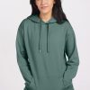 A person wearing an Avery Hoodie in Duck Green from Woolx stands against a neutral background, looking at the camera. Their dark hair falls loosely over their shoulders as their hands rest in the front pocket of the comfortable Merino wool garment. | Sarah is 5'8", wearing a size S