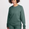 A person with curly hair smiles while wearing the Bailey Crewneck Sweatshirt in Duck Green, a sustainable piece from Woolx, along with matching pants. They are standing against a plain white background. | Tori is 5'7", wearing a size S