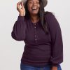 A smiling woman poses against a plain background, wearing the Woolx Charlotte Puff Sleeve Top in Deep Plum and jeans. Her long dark hair cascades down as she holds a light gray hat, exuding elegance and charm.