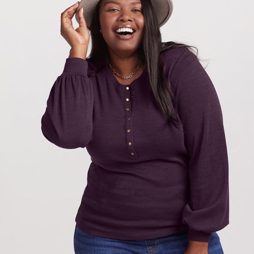 A smiling woman poses against a plain background, wearing the Woolx Charlotte Puff Sleeve Top in Deep Plum and jeans. Her long dark hair cascades down as she holds a light gray hat, exuding elegance and charm.