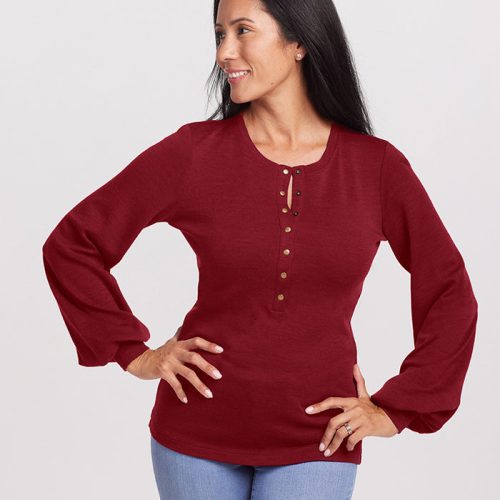 A person with long dark hair wears the Charlotte Puff Sleeve Top in Sweet Pepper, a luxurious piece crafted from Merino wool by Woolx. They pair it with blue jeans and stand confidently with one hand on their hip, smiling and looking to the side against a light gray background.