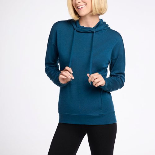 A person with short blonde hair is standing and smiling, dressed in a Woolx Callie Hoodie in Real Teal made from Australian Merino Wool and black leggings, holding onto the drawstrings of their hoodie. The background is plain white. | Georgia is 5'10", wearing a size XS