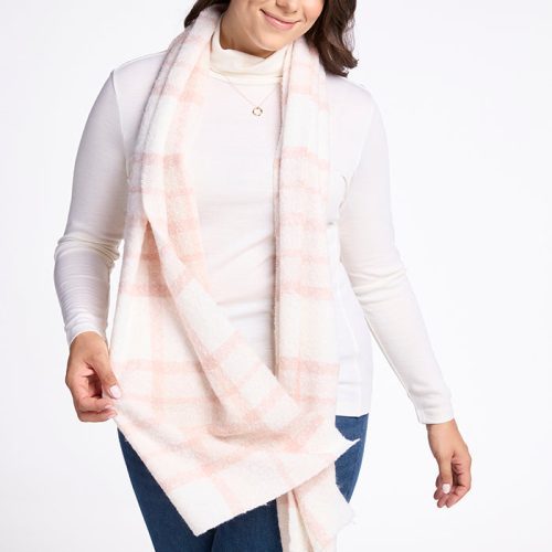 A person with shoulder-length dark hair is posing against a white background. They are wearing a long, mauve plaid scarf named the Bradie Scarf by Woolx, a white turtleneck, and blue jeans. They have a gentle smile and are holding the scarf with one hand.