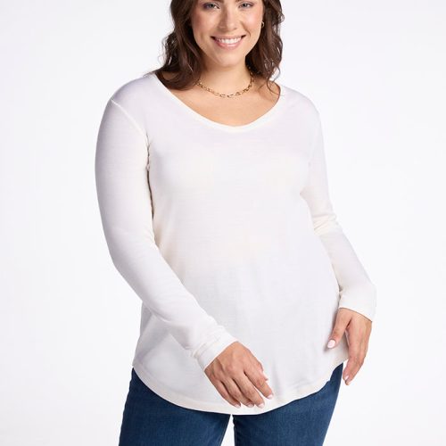 A person with long brown hair smiles while wearing the cozy Eva Tunic in Buttercream from Woolx and blue jeans. They stand against a plain white background, exuding a comfortable feel.