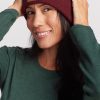 A person with long dark hair wearing a green long-sleeve shirt and a Woolx Baylor Beanie in Cranberry Melange is smiling and holding the sides of the beanie. The Merino wool beanie features a small rectangular patch with a simple line drawing of a bird. The background is plain and light-colored.