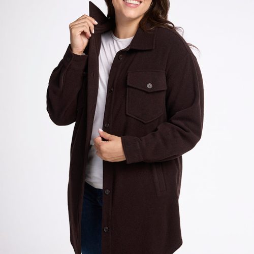 A smiling person with long dark hair wears the Sutton Shacket in French Roast from Woolx over a white shirt and blue jeans. They stand against a plain white background, confidently holding the collar of their shacket with one hand.