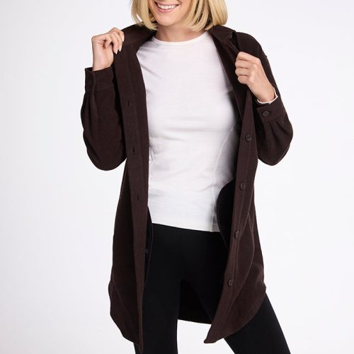 A person with shoulder-length blonde hair smiles while wearing a white shirt, black pants, and an open Sutton Shacket - French Roast by Woolx. They are holding the shacket’s lapels and standing against a plain white background, enjoying its breathable and moisture-wicking comfort.