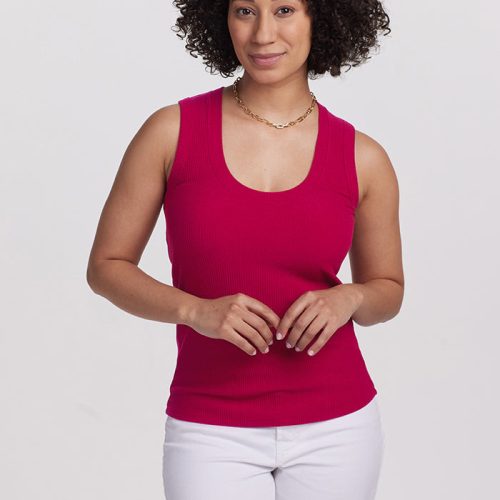 A person with curly hair smiles at the camera, wearing a vibrant Viva Magenta Sloane Ribbed Tank by Woolx and white pants offering a versatile fit. They stand against a plain, light gray background.