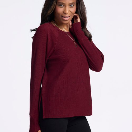 A woman is standing against a plain background, smiling and looking slightly to the side. She is wearing the Ainsley Sweater in Cranberry Melange by Woolx with black pants. She has long, dark hair. One hand is touching her sweater near her face, and the other is by her side. | Keesha is 5'9.5", wearing a size S