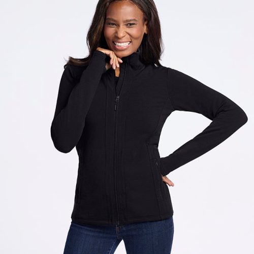 A woman with long hair is smiling and posing against a plain white background. She is wearing the Finley Zip Up Sweatshirt in black from Woolx, made of luxurious Australian merino wool, paired with blue jeans. With her left hand on her hip and her right hand touching her face, she effortlessly showcases the flattering look provided by the no-hood design of the sweatshirt.