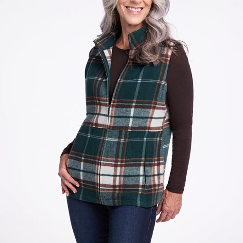 A smiling woman with long, wavy gray hair is wearing the Woolx Colbie Vest in Expresso Plaid over a dark long-sleeved shirt and blue jeans. She stands against a plain white background with one hand resting on her hip. | Tam is 5’8.5”, wearing a size S