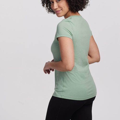 A person with curly hair is smiling and looking over their shoulder, wearing the ultra-soft Woolx Addie Short Sleeve Crew in Basil, combined with black pants. The background is plain and light, enhancing the fresh, odor-free vibe.