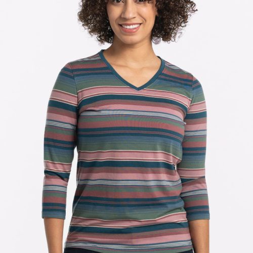 A person with curly hair smiles warmly, wearing the Woolx Elena V Neck in Adirondack 2.0 Stripe, crafted from lightweight fabric in shades of pink, green, and blue. Paired with dark jeans, they stand against a plain, light background. | Tori is 5'7", wearing a size S