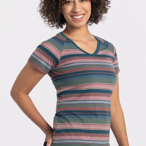 A person with curly hair smiles while wearing the vibrant, short-sleeved Mia V Neck - Adirondack 2.0 Stripe shirt by Woolx, paired with jeans. They are standing against a plain white background. | Tori is 5'7", wearing a size S