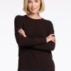 A person with short blonde hair wearing a Woolx Ainsley Sweater in French Roast and blue jeans stands facing the camera with arms crossed, smiling. The background is white. | Georgia is 5'10", wearing a size XS