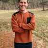 An outdoor enthusiast stands on a grassy, pine needle-covered field, smiling with arms crossed. They're wearing an abrasion-resistant Woolx Andes Crewneck Top in copper with black shoulder accents and dark pants. Tall trees and a forest form the scenic background.