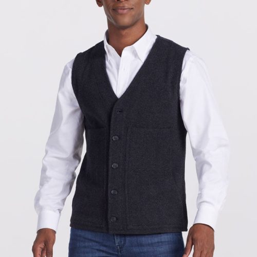 A person wearing a white dress shirt, a dark "Baker Vest OLD" from Woolx in carbon black, and blue jeans poses against a plain white background. The vest is buttoned down, providing insulation even when wet. The person smiles slightly with their hands relaxed by their sides.