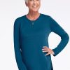 A mature woman with short white hair smiles confidently in a Real Teal Blair Tunic from Woolx paired with black pants. With one hand on her hip, she exudes elegance, appreciating the tunic's odor-resistant, lightweight warmth. The plain white background accentuates her poised presence.