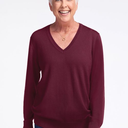 An older person with short white hair is smiling, wearing the Francesca Sweater in Cabernet Crush by Woolx and blue jeans, standing against a plain white background. | Kathy is 5'9", wearing a size S