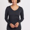 A person with curly hair is smiling and wearing the Woolx Eva Tunic in Carbon Stripe, a long-sleeve Merino wool V-neck top. They are standing against a plain, light-colored background, exuding street style with their hands gently clasped in front. | Tori is 5'7", wearing a size S