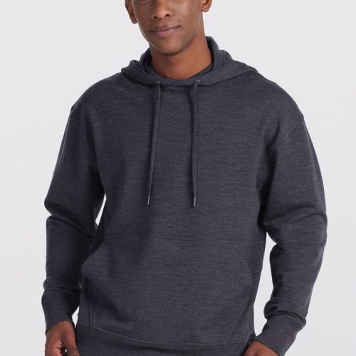 A person stands against a plain background, wearing the Woolx Chase Hoodie in Pebble Grey Melange with drawstrings and blue jeans. The individual is looking at the camera with a slight smile, their hands relaxed at their sides with thumbs in their pockets.