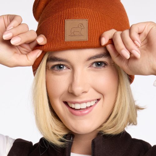 A person wearing a Woolx Baylor Beanie in Cinnamon Stick, smiling and holding the sides of their orange merino wool hat. Their blonde hair complements the dark vest over a white shirt against the plain white background.