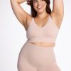 Wearing the Marley Shapewear Shorts in Sand by Woolx, a woman smiles confidently as she highlights the gentle compression of her outfit. One arm is raised while her other hand rests on her head against a plain white background. | Juliana is 5'10", wearing a size XL