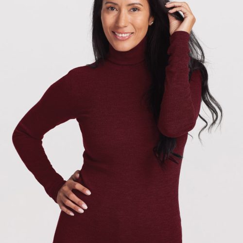 A person with long dark hair smiles while wearing the Woolx Sage Ribbed Turtleneck in Cranberry Melange and blue jeans. They are standing against a plain white background, with one hand on their hip and the other touching their hair. | Denia is 5'8", wearing a size S