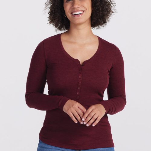 A smiling woman with curly hair wears the Reese Ribbed Henley in Cranberry Melange by Woolx, paired with blue jeans. Made from Merino wool, her top features moisture-wicking properties. She stands against a plain white background, looking directly at the camera with her hands clasped in front. | Tori is 5'7", wearing a size S