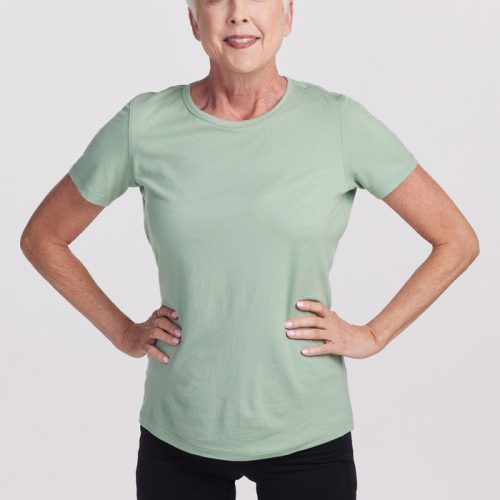 An older woman with short gray hair and a confident smile stands with hands on her hips. She's wearing a lightweight Liza Crew Neck tee in Basil from Woolx, paired with black shorts, posing against a plain white background.