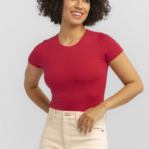 A person with curly hair smiles in ultimate comfort, wearing a Viva Magenta Gracie Bodysuit from Woolx. They stand against a plain, light gray background, exuding a relaxed vibe perfect for any summer wardrobe.