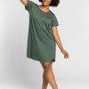 A person with curly hair smiles while posing in a Duck Green Desi Nightgown OLD by Woolx. They stand barefoot against a plain, light gray background, gracefully raising one arm above their head.