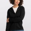 A person with curly hair is wearing a Woolx Cubby Hooded Sweatshirt in black with heavy-duty zippers over a white shirt and blue jeans. They are standing against a plain light-colored background, with one arm crossing their body and the other hanging by their side. | Tori is 5'7", wearing a size S